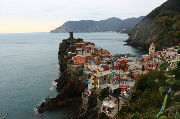 Vista de cima do vilarejo de Cinque Terre,  região da Ligúria na Italia, com suas falesias rochosas e casas em tons de amarelo ensolarado, laranja  queimado e rosa suave, contrastando com as montanhas esverdeadas e o mar nos tons de turquesa, azul cobalto e verde esmeralda. Créditos da foto: Partha Samanta - Unsplash 